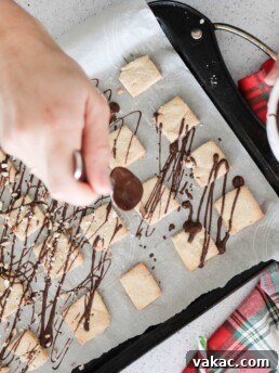 A stack of classic shortbread squares, some with chocolate drizzle, on a rustic wooden board.