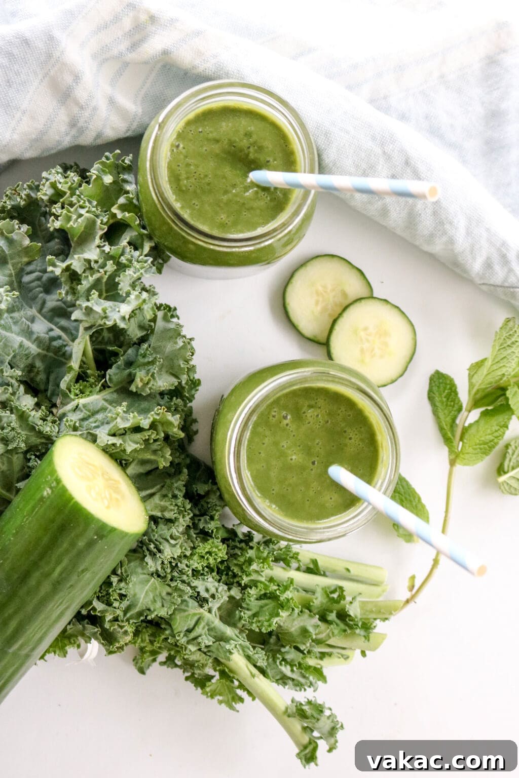 Two mason jars with straws filled with vibrant green juice, surrounded by fresh kale and cucumber slices on a rustic surface.