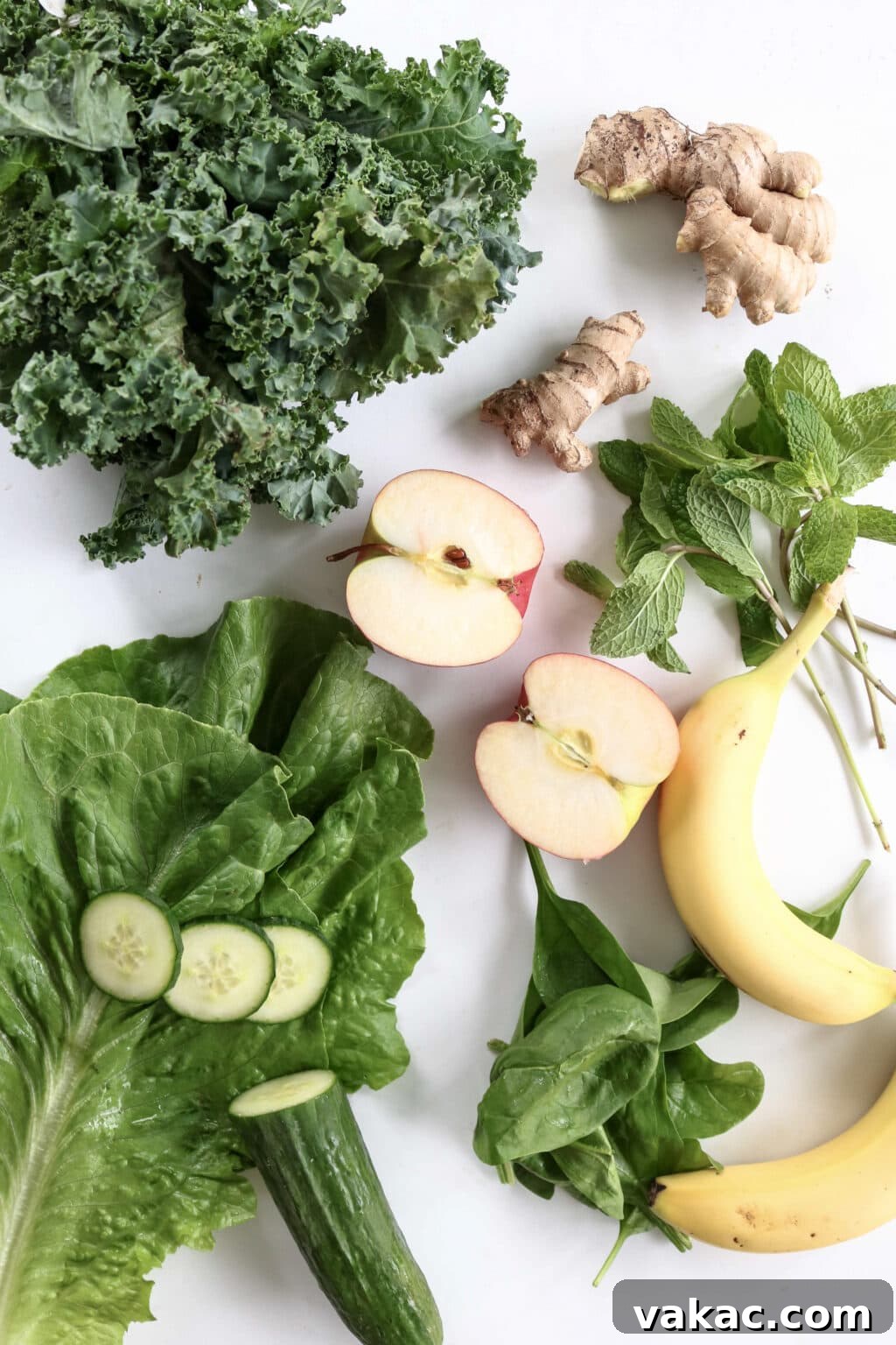 An inviting overhead shot displaying all the fresh ingredients for the green juice: vibrant curly kale, two pieces of fresh ginger, a crisp apple cut in half, fragrant mint leaves, a ripe banana, fresh romaine lettuce, and a hydrating cucumber.