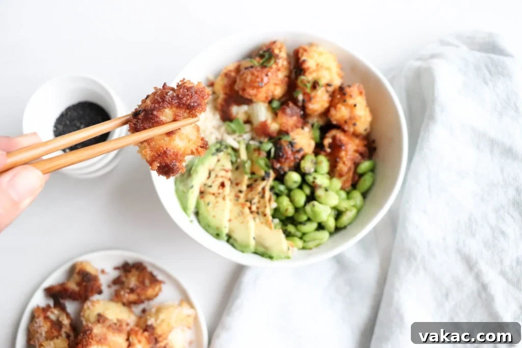 Golden Honey Garlic Cauliflower Power Bowl 2 A vibrant Crispy Baked Cauliflower Bowl resting on a clean white surface, accompanied by a neatly folded white kitchen towel. The bowl is generously filled with perfectly cooked rice, creamy avocado slices, bright green edamame, and the star of the show: golden-brown, crispy baked cauliflower florets.