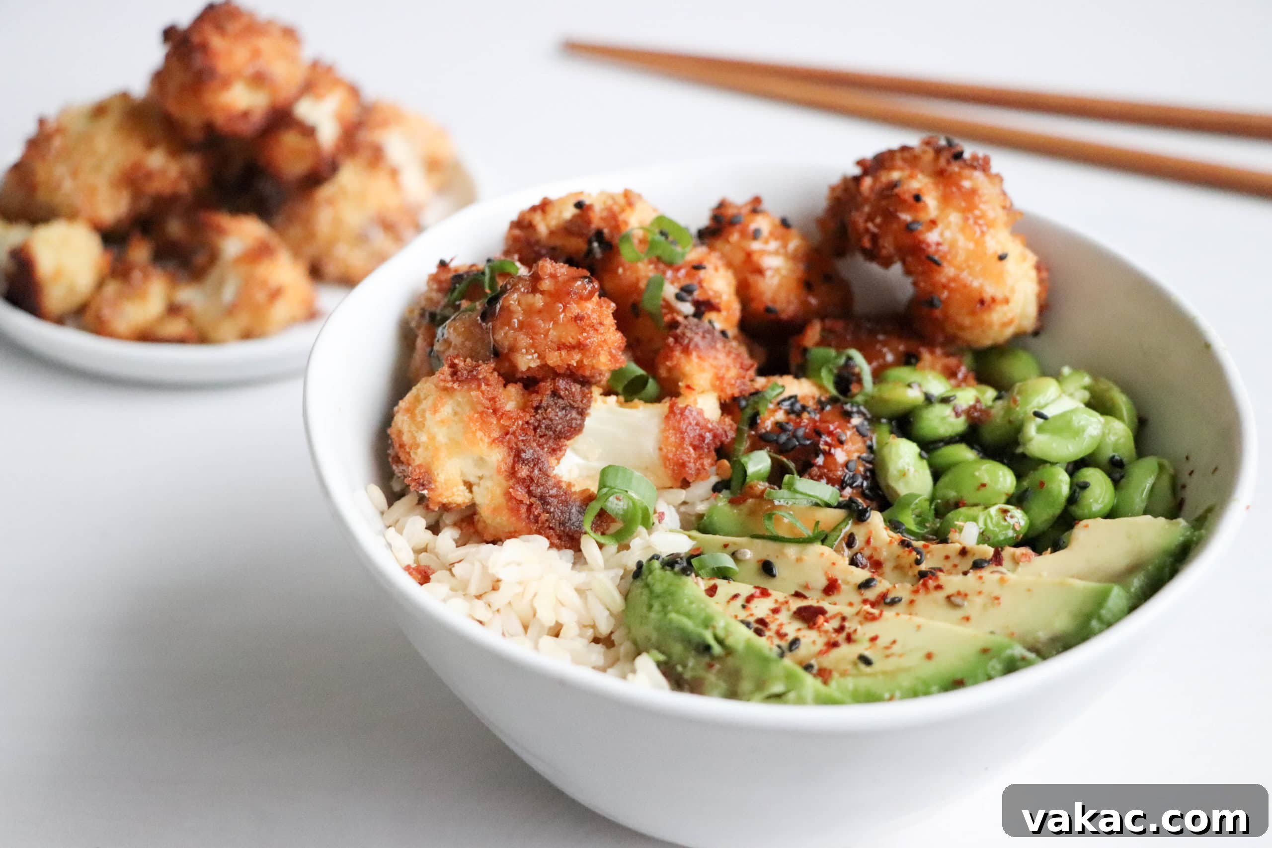 Golden Honey Garlic Cauliflower Power Bowl 4 A close-up shot of a white bowl overflowing with a Crispy Baked Cauliflower Bowl, featuring rice, avocado, edamame, and the signature crispy cauliflower. A plate of additional crispy cauliflower florets sits beside it on a clean white surface.