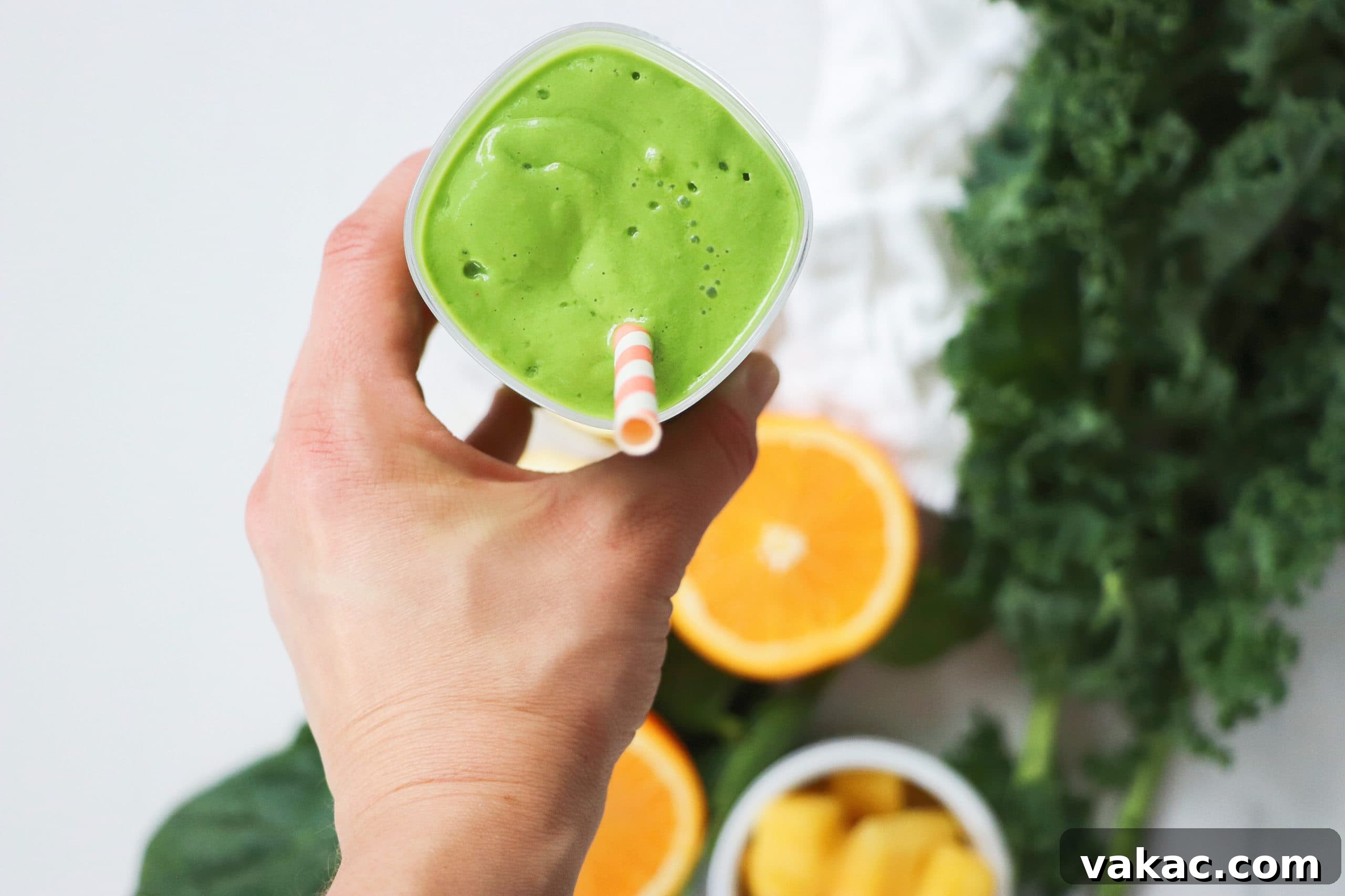 Quick Prenatal Vitality Smoothie 3 The author, Lindsay Pleskot, proudly holding a pregnancy green smoothie in a glass with a striped straw. The background showcases a white board adorned with fresh oranges, mango, and leafy greens, illustrating the smoothie's natural ingredients.