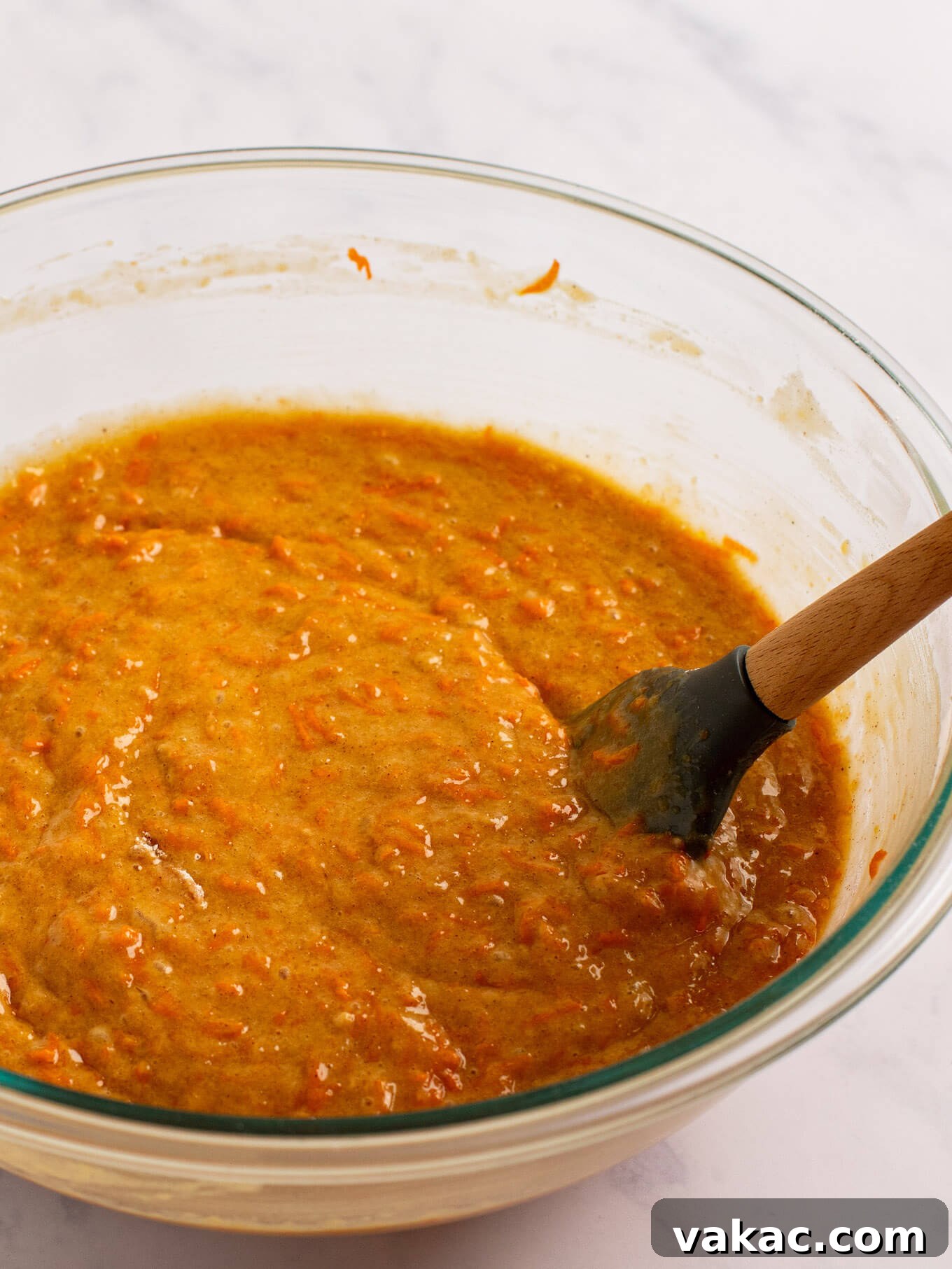Carrot cake batter in a glass mixing bowl. A rubber spatula rests on the side of the bowl.