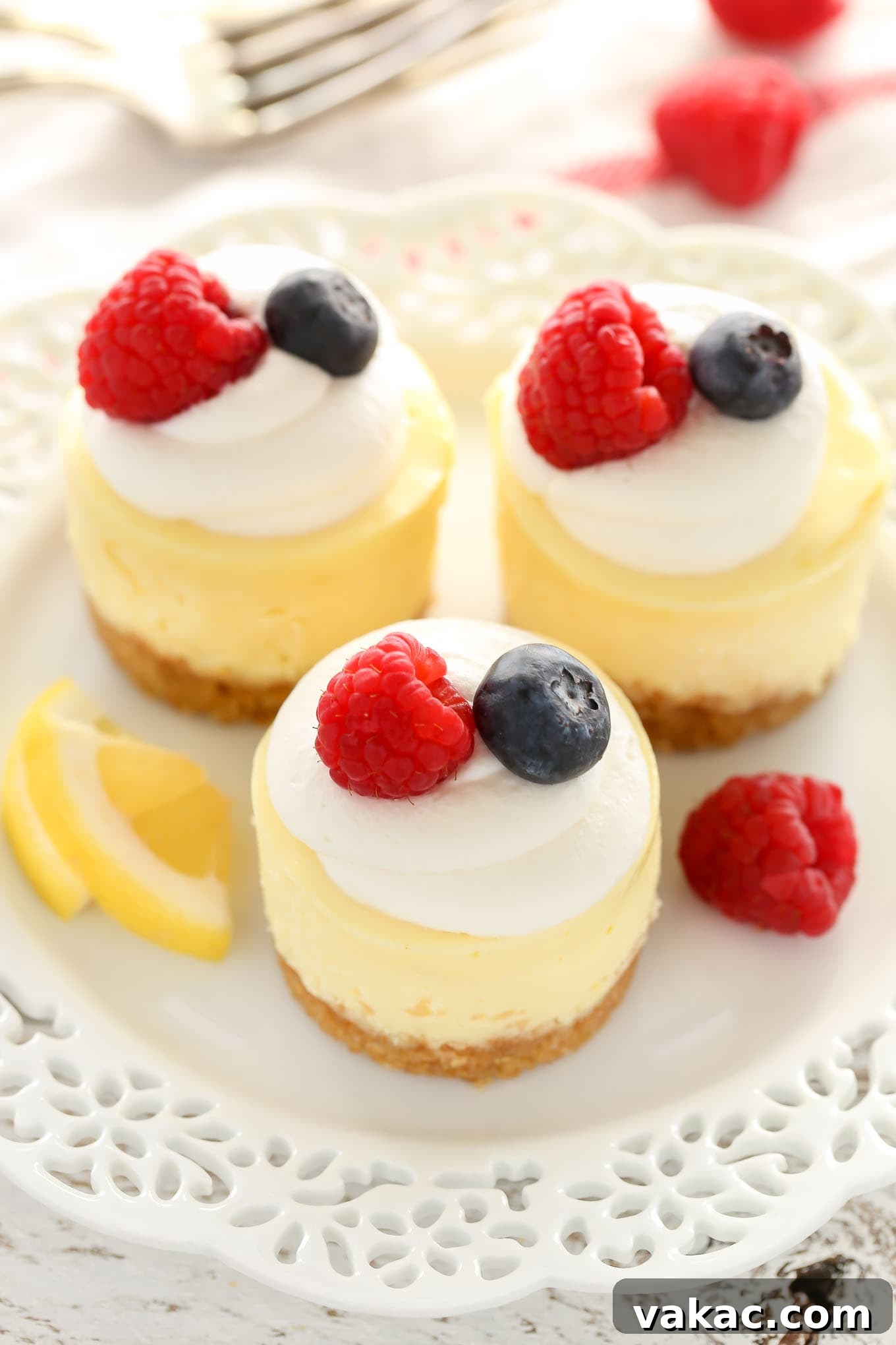 Sunshine Lemon Cheesecake Bites 2 Overhead view of three mini lemon cheesecakes garnished with whipped cream and berries on a white plate. A fork rests in the background.