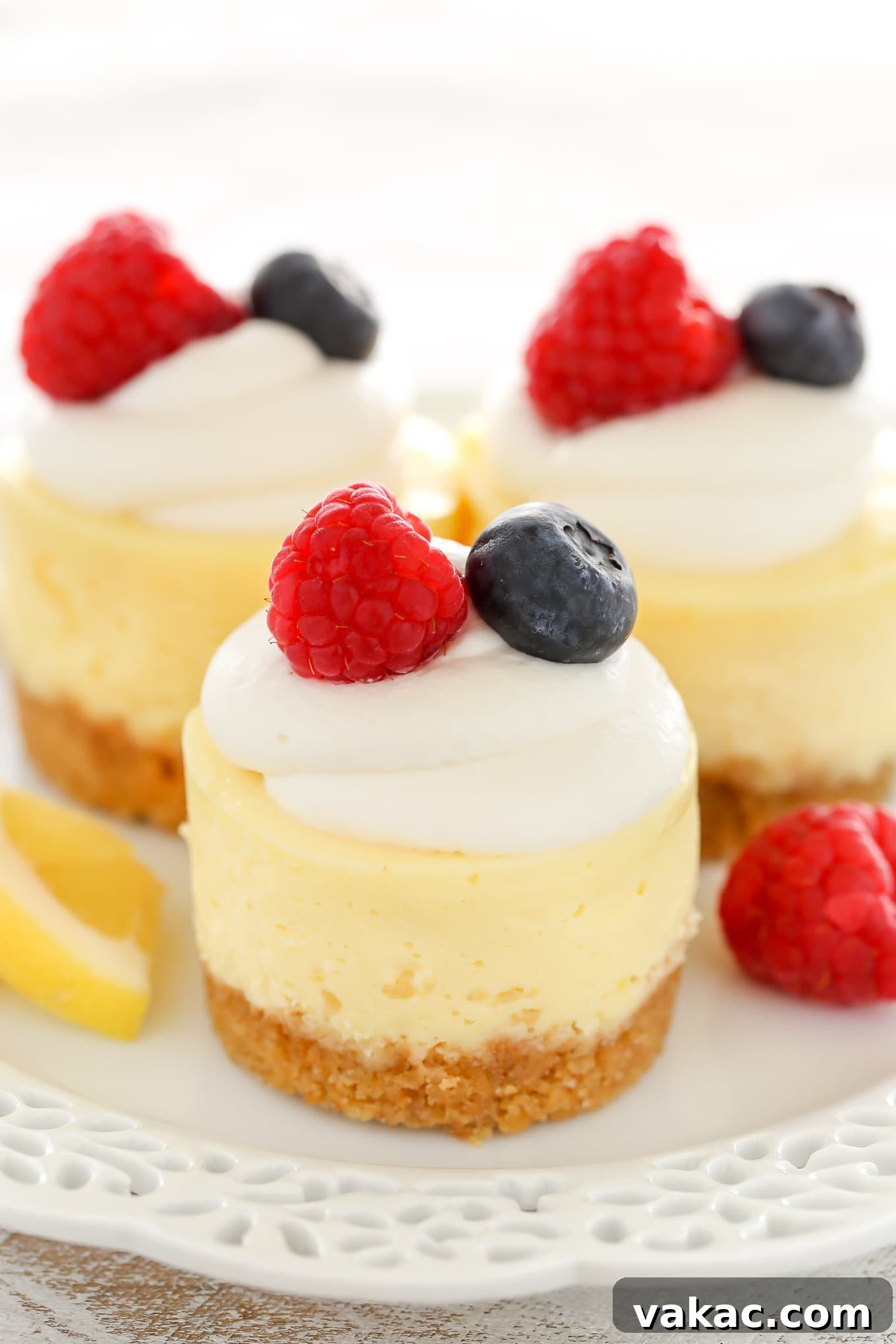Sunshine Lemon Cheesecake Bites 3 Three mini cheesecakes with a graham cracker crust on a white plate. Each mini cheesecake is garnished with whipped cream and fresh berries.