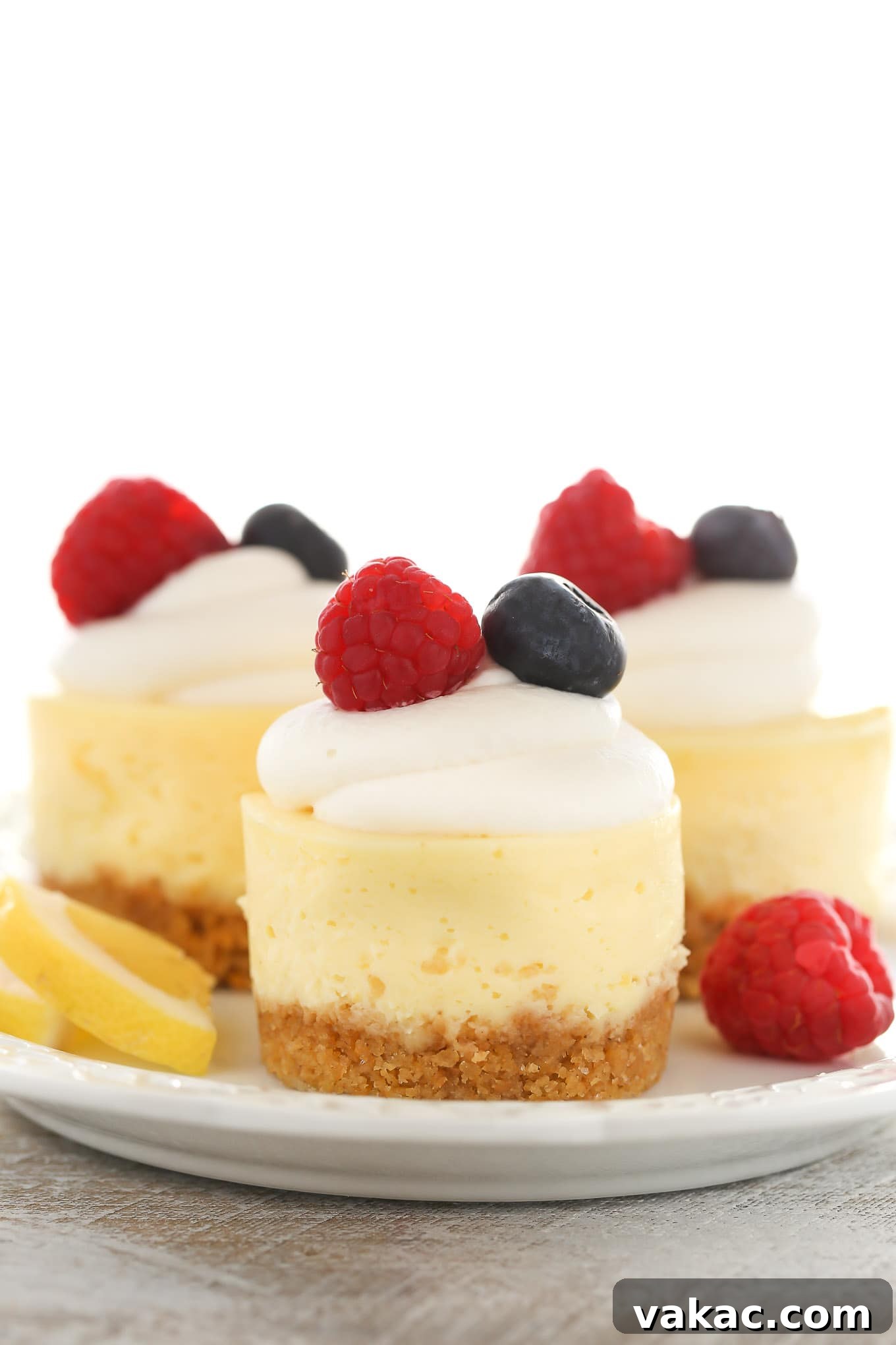 Sunshine Lemon Cheesecake Bites 4 A side view of three lemon cheesecake bites on a white plate. Each is topped with whipped cream and fresh berries.