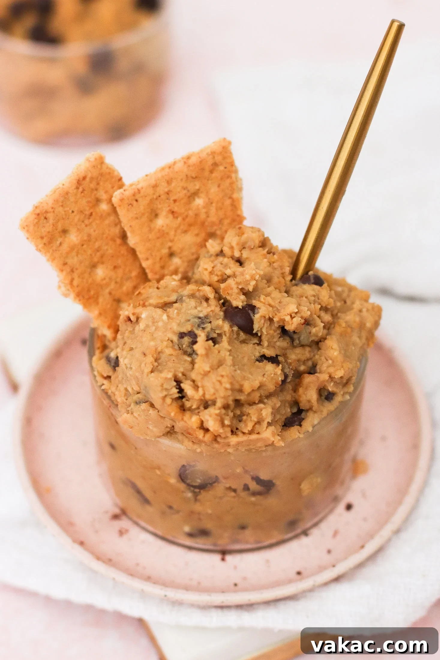 Guilt-Free High-Protein Chickpea Cookie Dough 2 A glass cup filled with chocolate chip cookie dough, garnished with two rectangular graham crackers and a gold spoon, sits on a small pink plate atop a white napkin, highlighting a delicious and healthy snack.