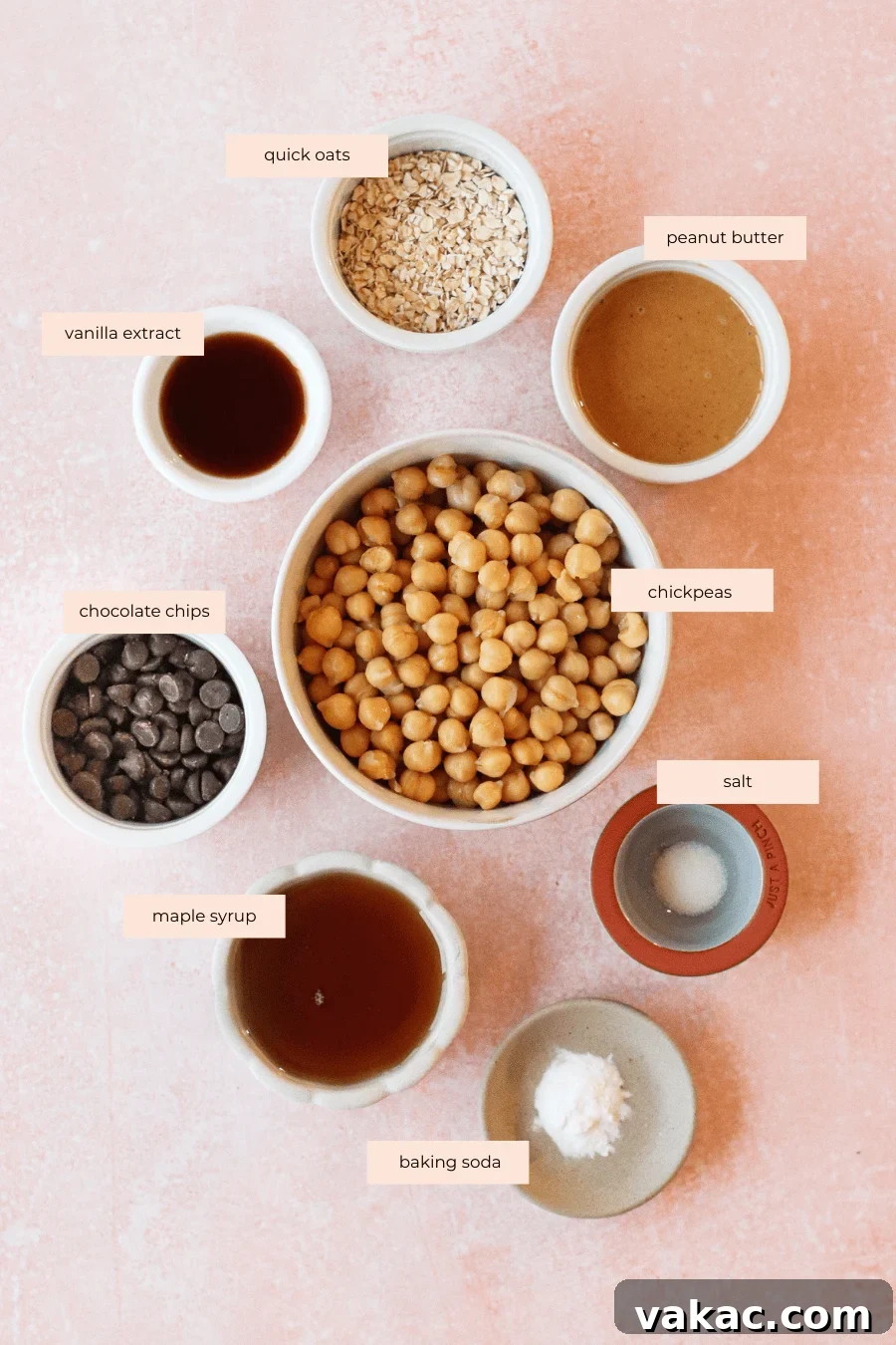 Guilt-Free High-Protein Chickpea Cookie Dough 3 Eight bowls on a pink surface, each labeled: quick oats, peanut butter, chickpeas, chocolate chips, vanilla extract, maple syrup, salt, and baking soda—ingredients for edible chickpea cookie dough.