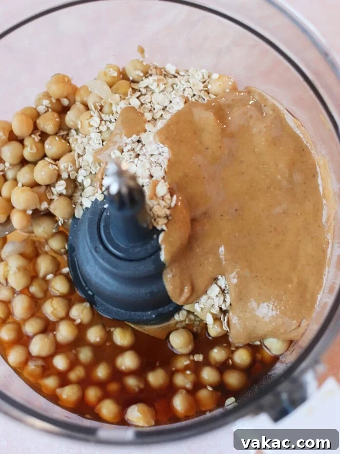 Guilt-Free High-Protein Chickpea Cookie Dough 4 A food processor containing chickpeas, rolled oats, almond butter, and a brown liquid ingredient, viewed from above before blending for chickpea cookie dough.