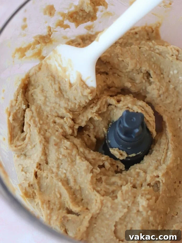 Guilt-Free High-Protein Chickpea Cookie Dough 5 A close-up of thick, beige chickpea cookie dough being mixed in a food processor with a white spatula, demonstrating its creamy texture.