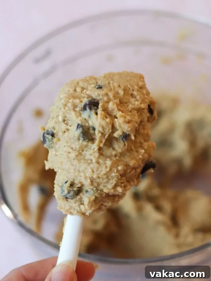 Guilt-Free High-Protein Chickpea Cookie Dough 6 A close-up of a hand holding a spatula with chocolate chip cookie dough above a glass mixing bowl filled with more dough, ready for chilling.