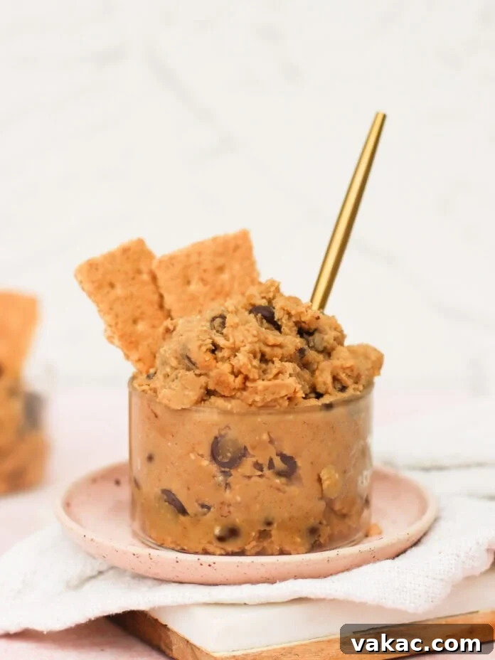 Guilt-Free High-Protein Chickpea Cookie Dough 7 A small glass jar filled with chocolate chip cookie dough, topped with two graham cracker pieces and a gold spoon, sits on a pink plate with a beige napkin, ready to be enjoyed.