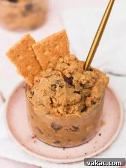 A glass cup filled with chocolate chip cookie dough, garnished with two rectangular graham crackers and a gold spoon, sits on a small pink plate atop a white napkin.
