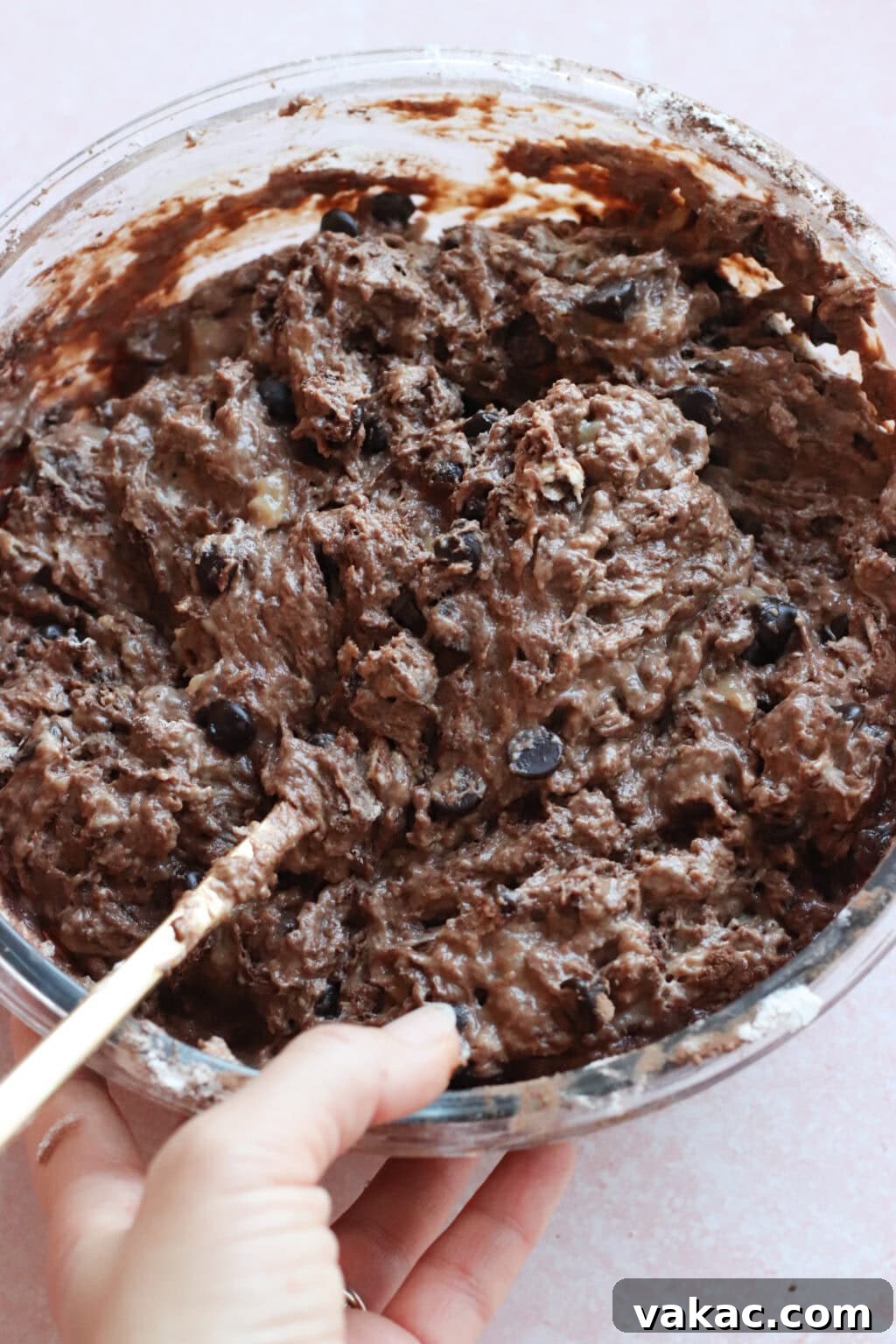 Velvety Moist Double Chocolate Muffins Naturally Packed with Protein 5 A hand holding a glass bowl filled with thick chocolate protein muffin batter, being mixed with a gold spoon.