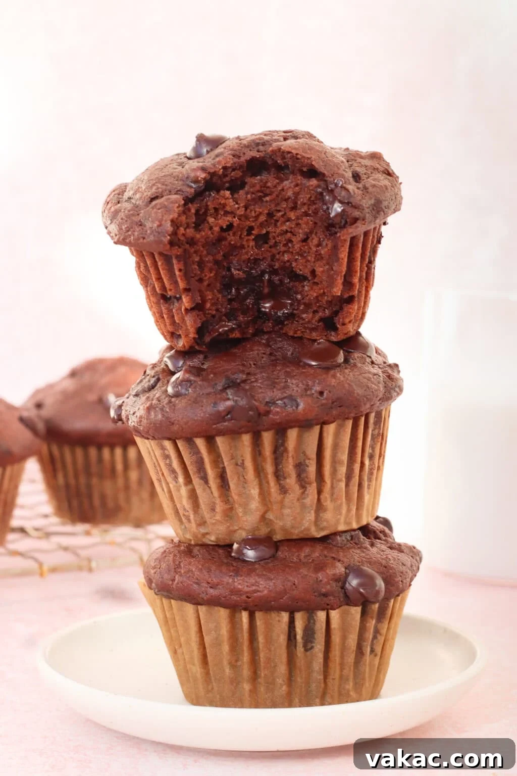 Velvety Moist Double Chocolate Muffins Naturally Packed with Protein 8 Three chocolate chip muffins are stacked on a white plate, with the top muffin showing a bite taken out. More muffins and a glass of milk are in the background, all set on a light pink surface.
