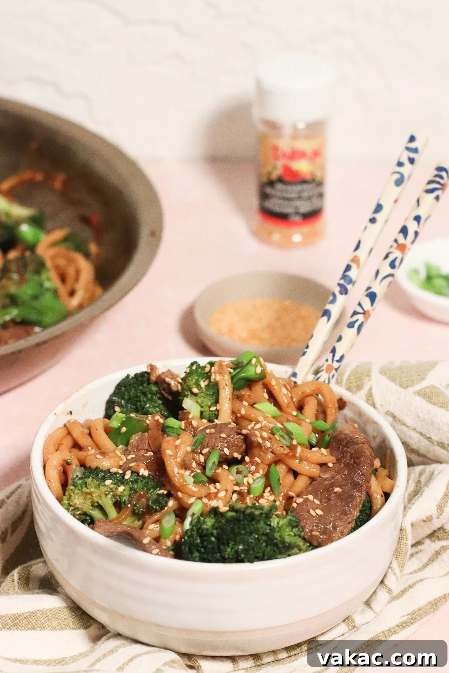 A bowl of stir-fried noodles with beef, broccoli, and onions, topped with sesame seeds and green onions. Chopsticks rest on the bowl, with a skillet, sesame seeds, and seasoning visible in the background.