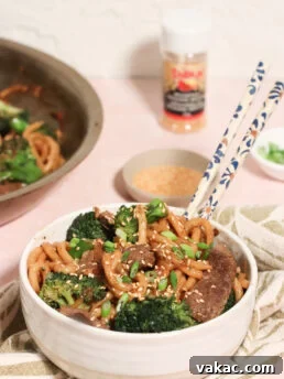 A bowl of stir-fried noodles with beef, broccoli, and onions, topped with sesame seeds and green onions. Chopsticks rest on the bowl, with a skillet, sesame seeds, and seasoning visible in the background.