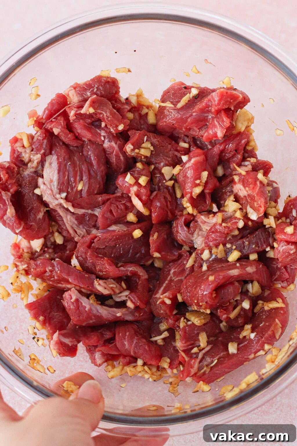 A clear glass bowl filled with sliced raw beef marinated with chopped garlic and ginger, held by a hand against a light pink background.