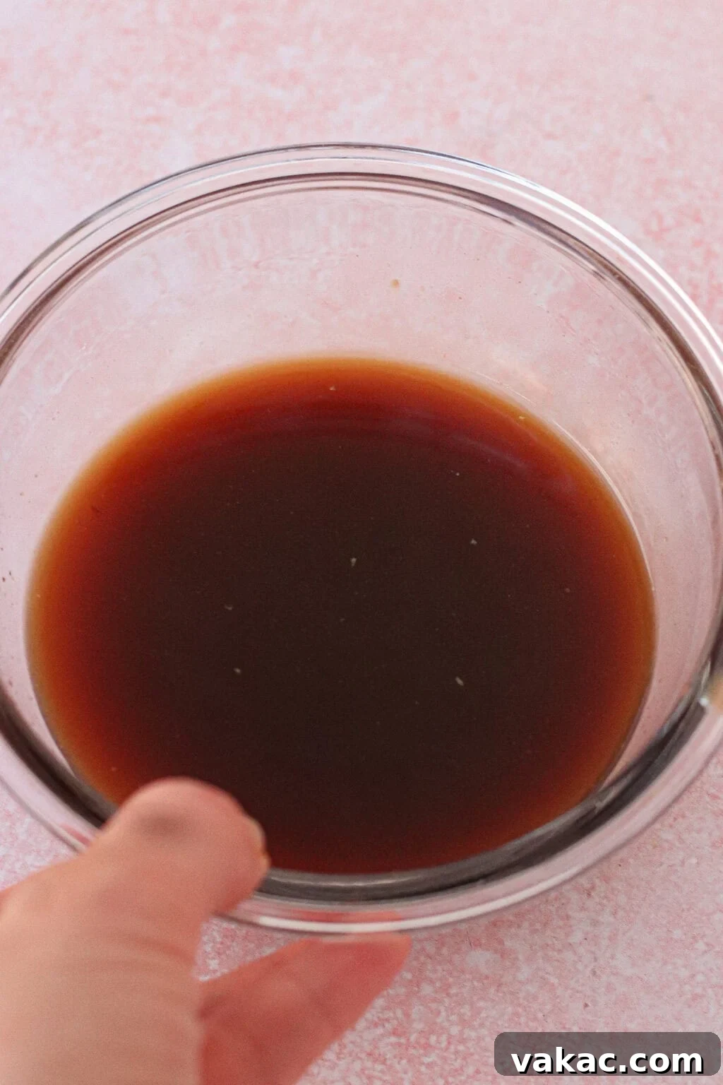 A hand holds a clear glass bowl filled with a dark brown liquid, set on a light pink surface.