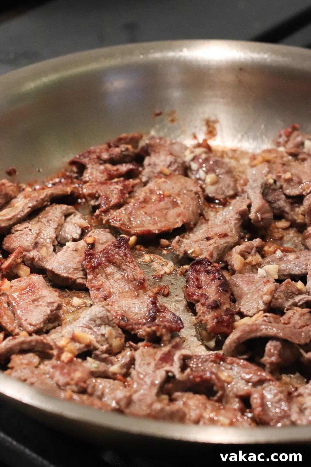 Slices of beef cooking and browning in a stainless steel skillet, with visible pieces of garlic and a caramelized surface on the meat.