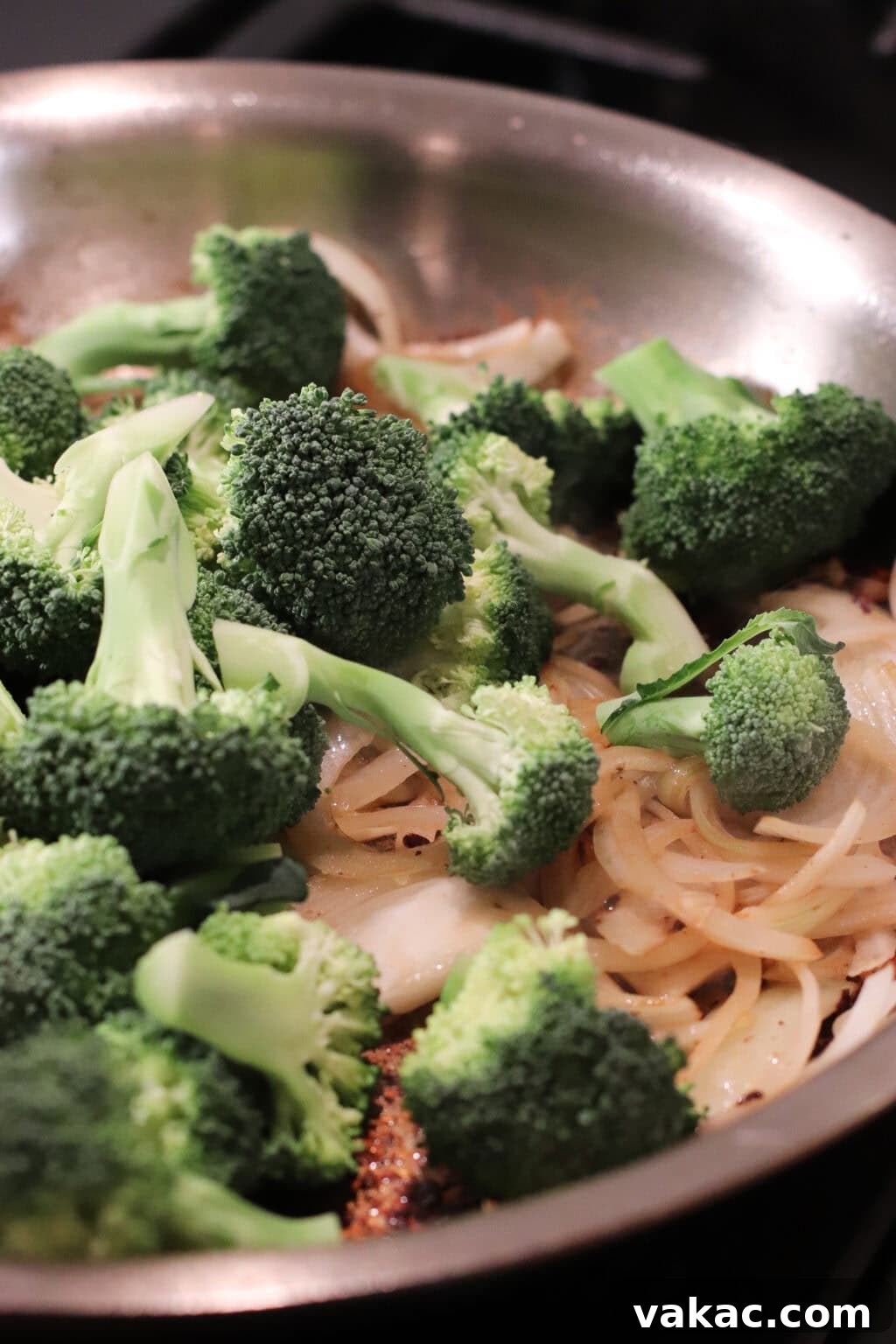 Broccoli florets and sliced onions cooking in a stainless steel pan on a stovetop, with some onions lightly browned and the broccoli bright green and fresh.