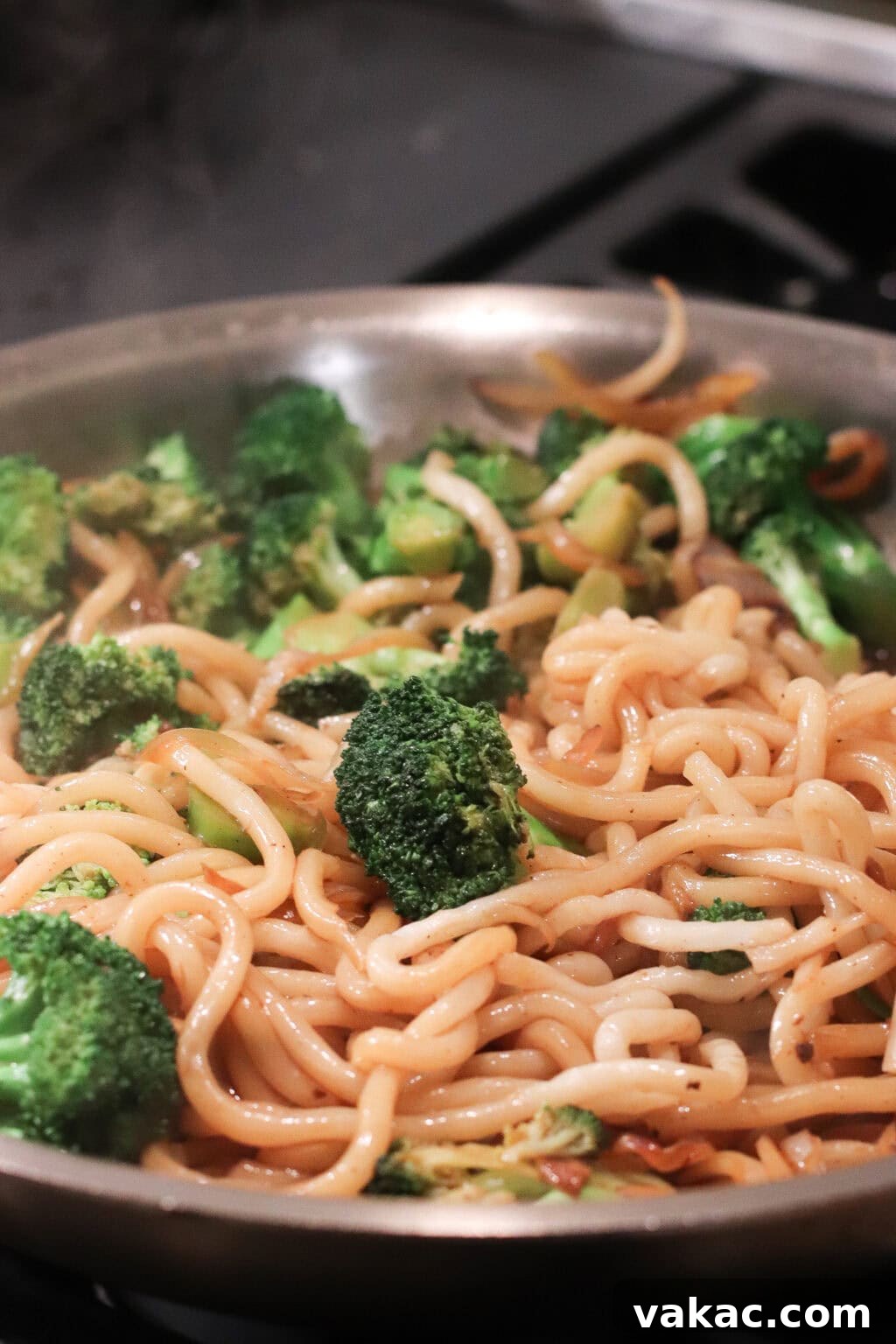 A close-up of a pan filled with stir-fried noodles, broccoli florets, and vegetables, all lightly coated in sauce. Steam rises from the hot dish, indicating it has just been cooked.