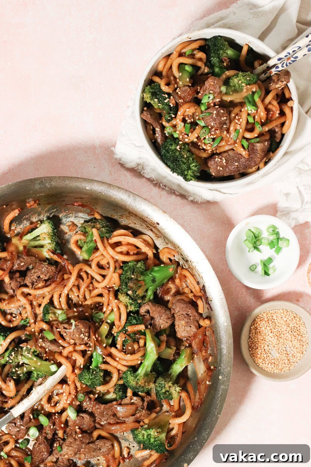 A skillet with beef, broccoli, and noodles in a savory sauce sits beside a bowl of the same dish, garnished with green onions and sesame seeds. Small bowls of green onions and sesame seeds are nearby on a pink surface.