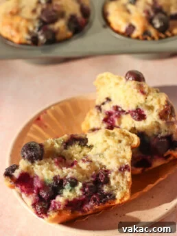 A blueberry muffin cut in half on a plate, showing a moist, fluffy texture with whole blueberries inside. In the background, more muffins are visible in a muffin pan.
