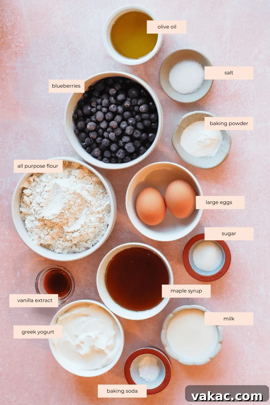 Wholesome Fluffy Blueberry Greek Yogurt Muffins 5g Protein 3 A flat lay of labeled baking ingredients for blueberry and Greek yogurt muffins on a pink background, including olive oil, salt, baking powder, blueberries, flour, eggs, sugar, maple syrup, vanilla extract, Greek yogurt, milk, and baking soda in bowls.