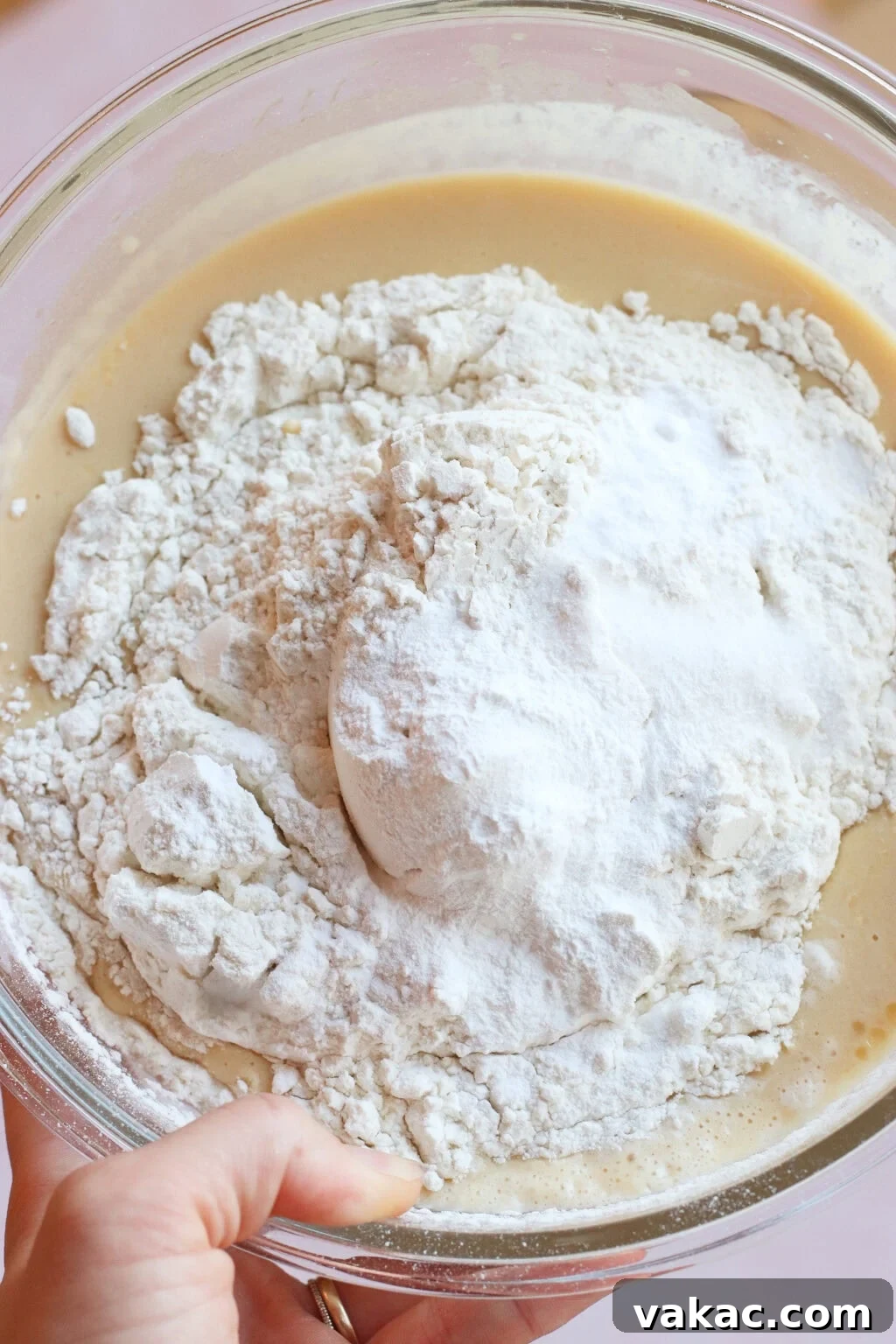 Wholesome Fluffy Blueberry Greek Yogurt Muffins 5g Protein 5 A close-up of a glass bowl containing a creamy batter topped with a mound of flour, held by a hand ready to mix the ingredients together.