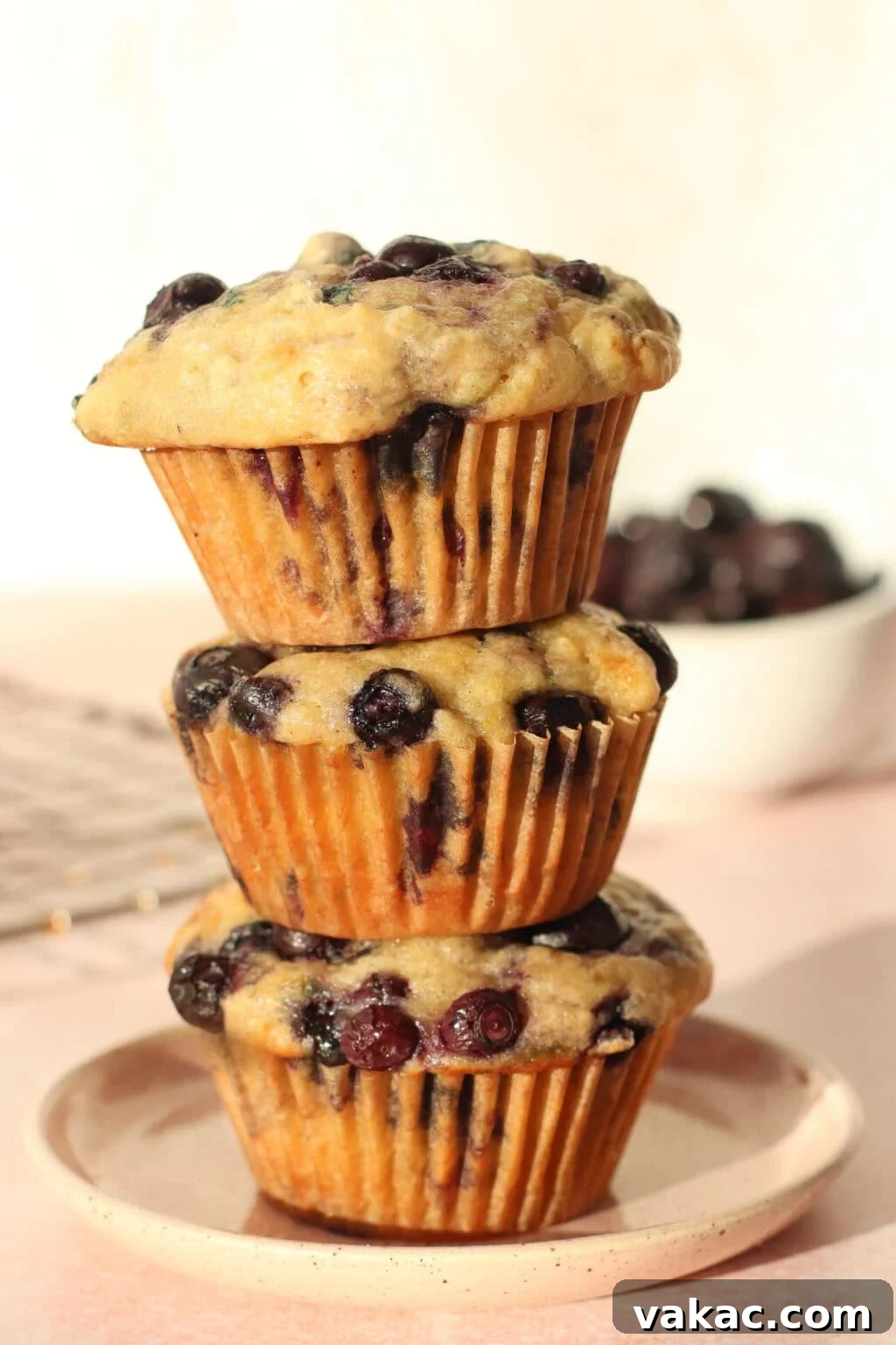 Wholesome Fluffy Blueberry Greek Yogurt Muffins 5g Protein 9 Three blueberry muffins are stacked on a round plate, with a bowl of blueberries and a cooling rack in the background, all set on a light pink surface.