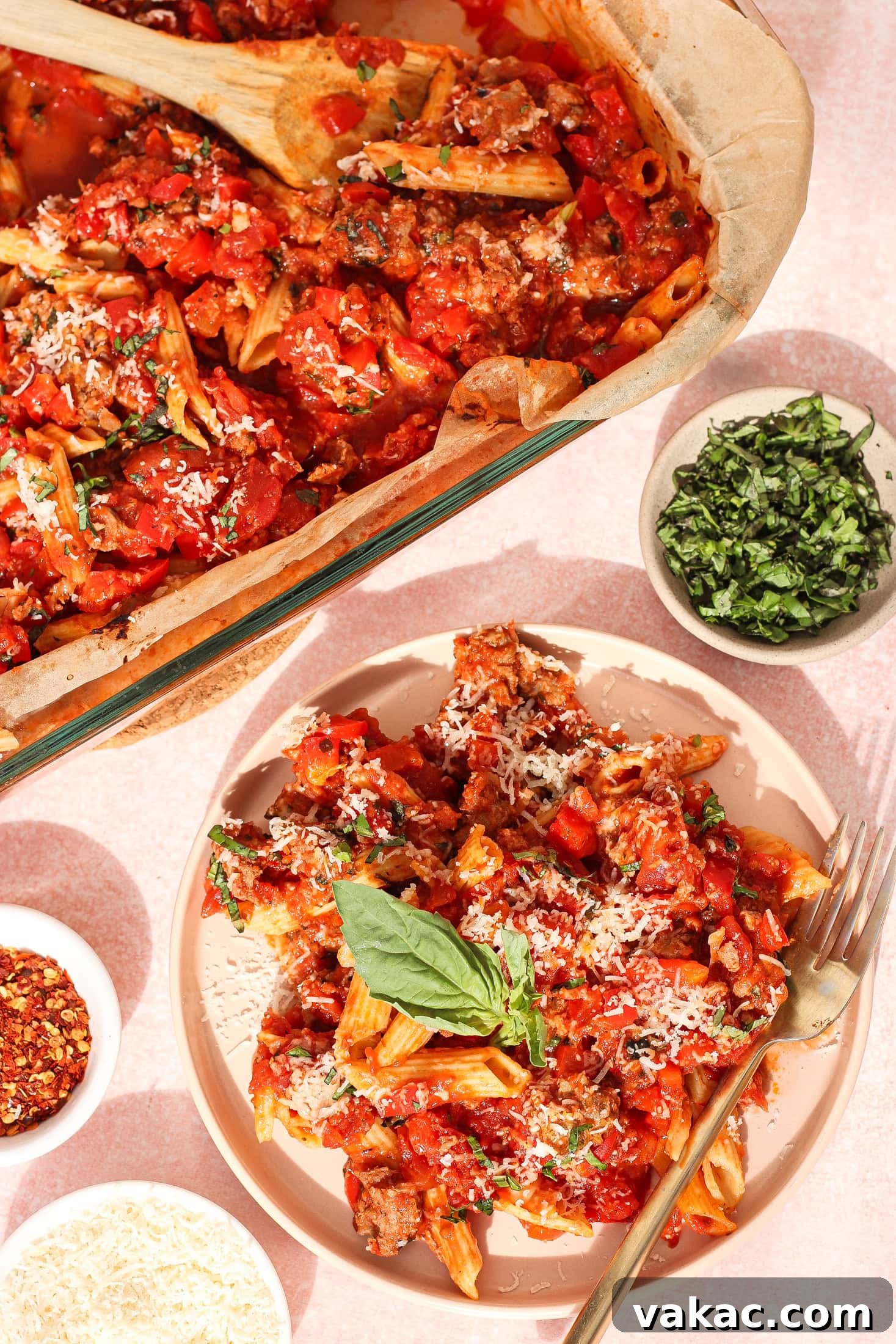 One Pan High Protein Sausage and Pepper Baked Pasta 2 A plate of baked pasta with tomato sauce, sausage, and grated cheese is garnished with fresh basil. Nearby are a baking dish of pasta, a bowl of chopped herbs, grated cheese, and red pepper flakes.