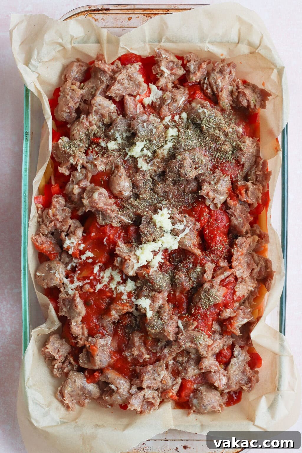 One Pan High Protein Sausage and Pepper Baked Pasta 4 A glass baking dish lined with parchment paper, filled with layers of tomato sauce, crumbled sausage, garlic, and herbs, ready to be baked.