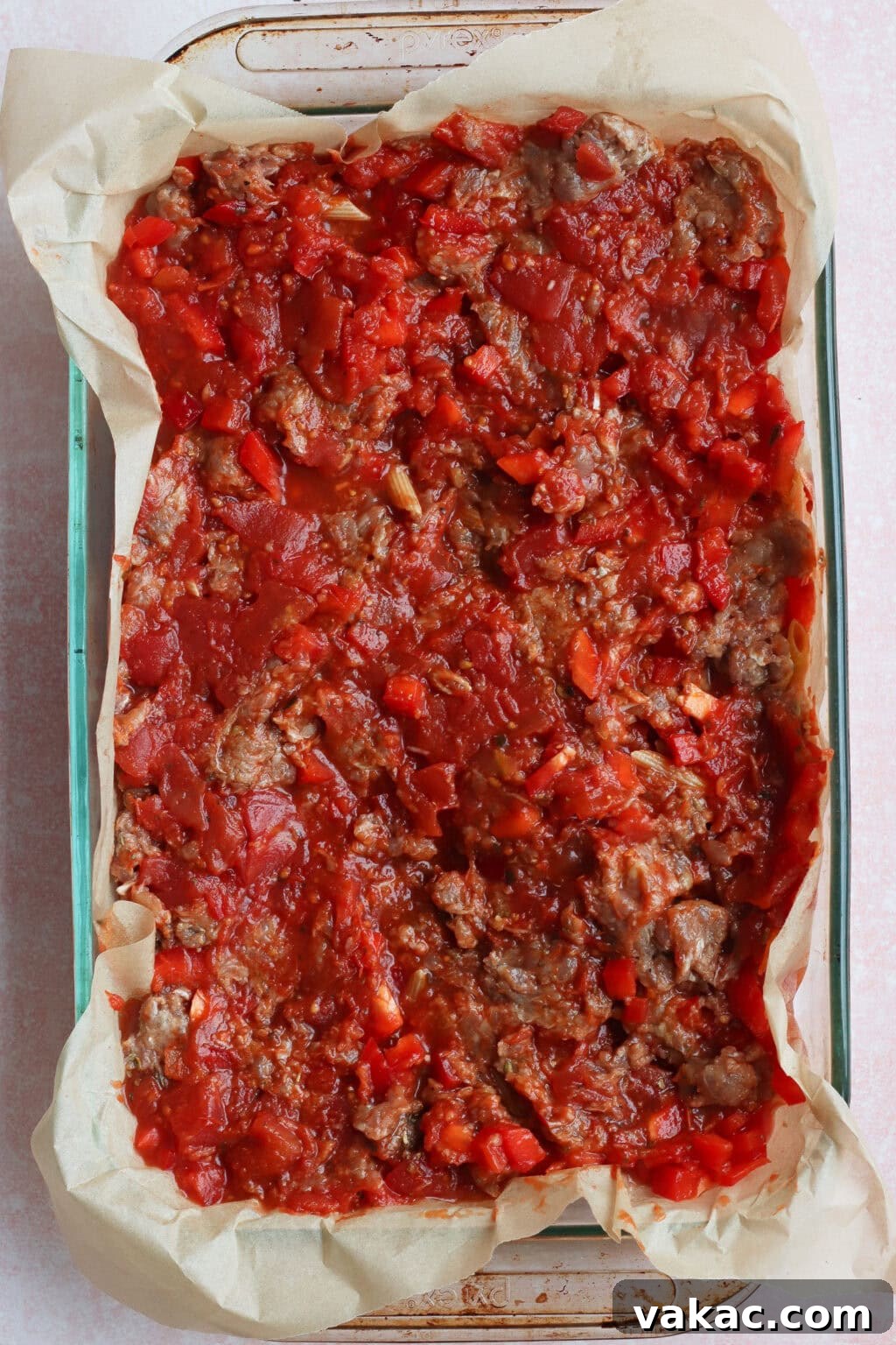 One Pan High Protein Sausage and Pepper Baked Pasta 5 A glass baking dish lined with parchment paper is filled with a chunky tomato and ground meat mixture, spread evenly and ready to be baked.