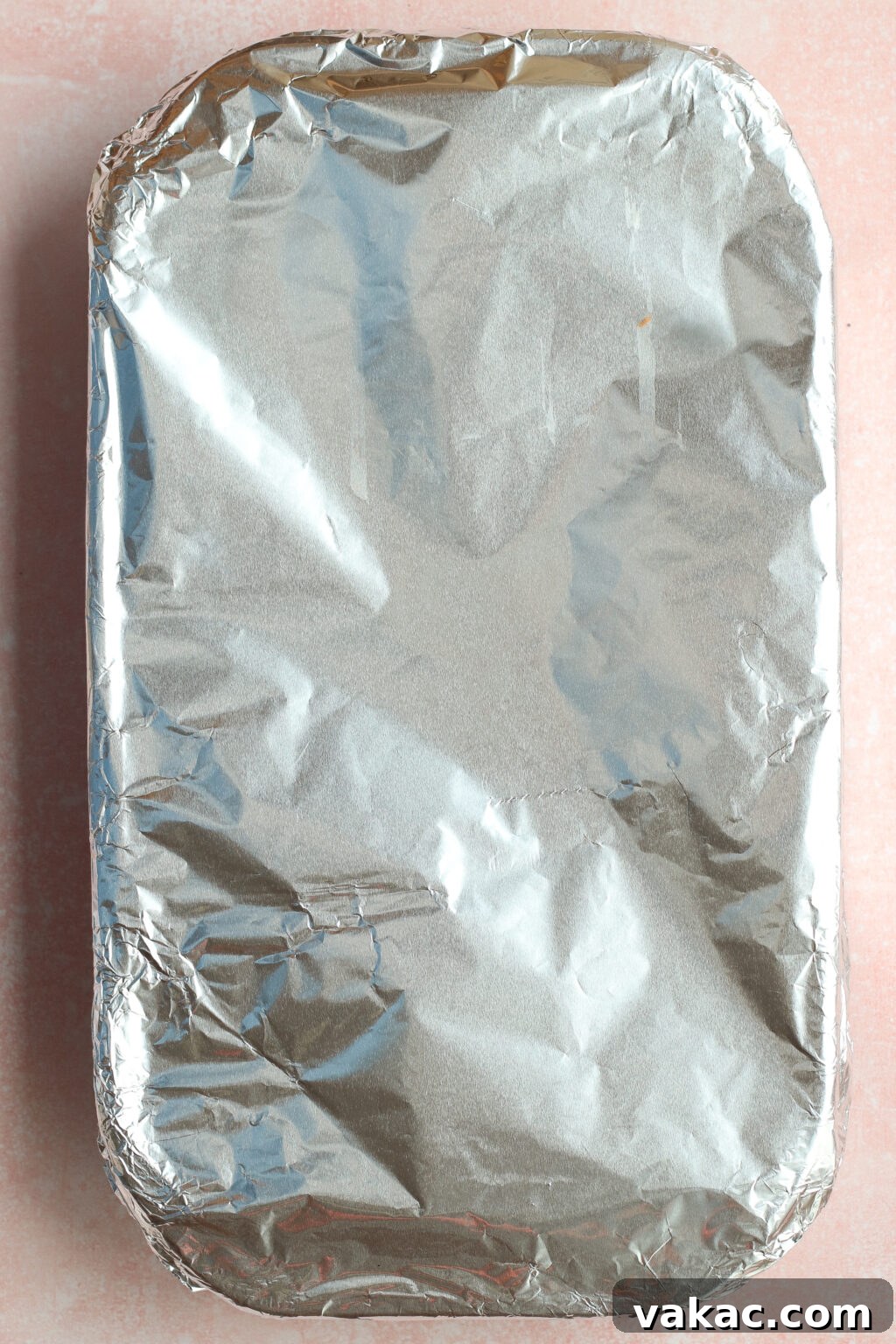 One Pan High Protein Sausage and Pepper Baked Pasta 6 A rectangular baking dish covered tightly with crinkled aluminum foil sits on a light pink surface.