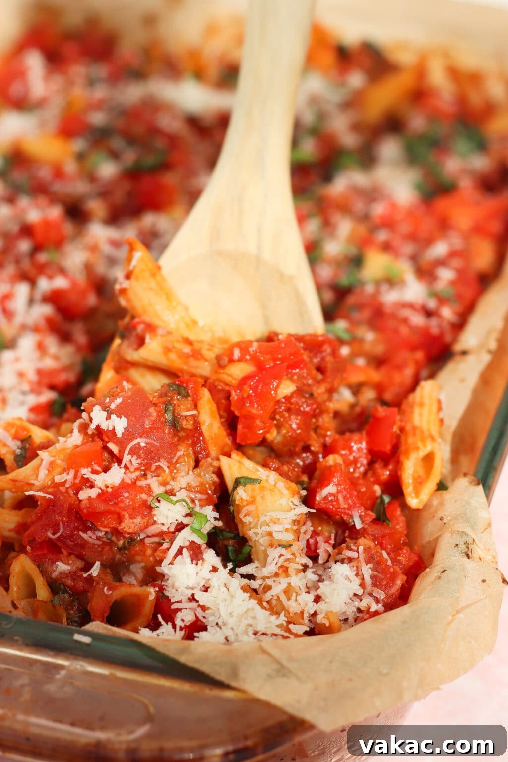 One Pan High Protein Sausage and Pepper Baked Pasta 7 A wooden spoon scoops baked penne pasta with tomato sauce and grated cheese from a parchment-lined baking dish. The pasta is garnished with fresh herbs and topped with shredded cheese.