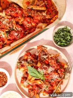 Baked pasta with tomato sauce, ground meat, and cheese is served on a plate with a basil leaf garnish. Nearby are bowls of grated cheese, chopped basil, and red pepper flakes. A fork rests on the plate.