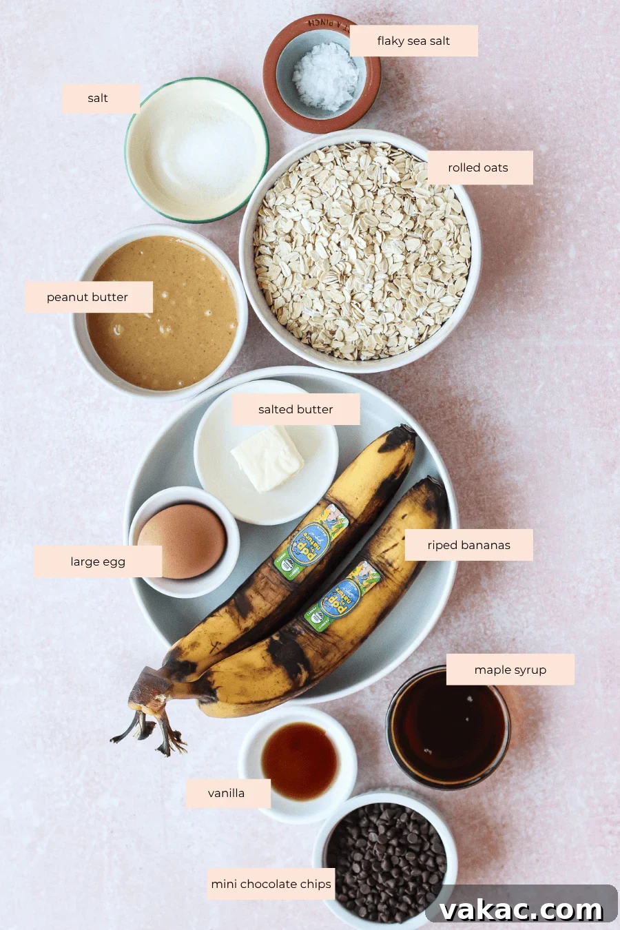 A flat lay of labeled baking ingredients: salt, flaky sea salt, rolled oats, peanut butter, salted butter, large egg, ripe bananas, maple syrup, vanilla, and mini chocolate chips on a light pink surface.