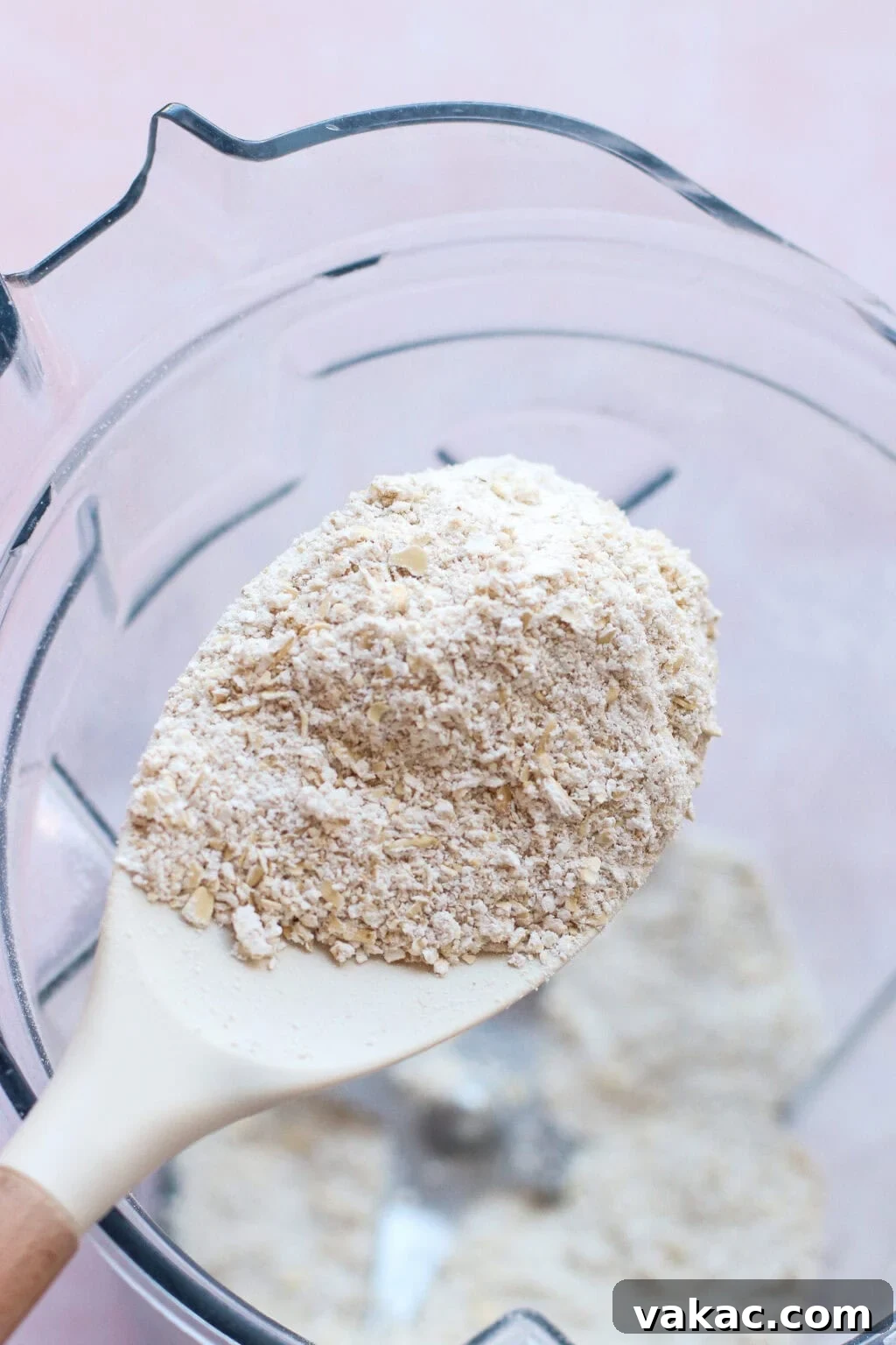 A close-up of a white spatula holding finely ground oat flour over a blender bowl filled with more oat flour. The background is soft pink, and the texture of the blended oats is visible.
