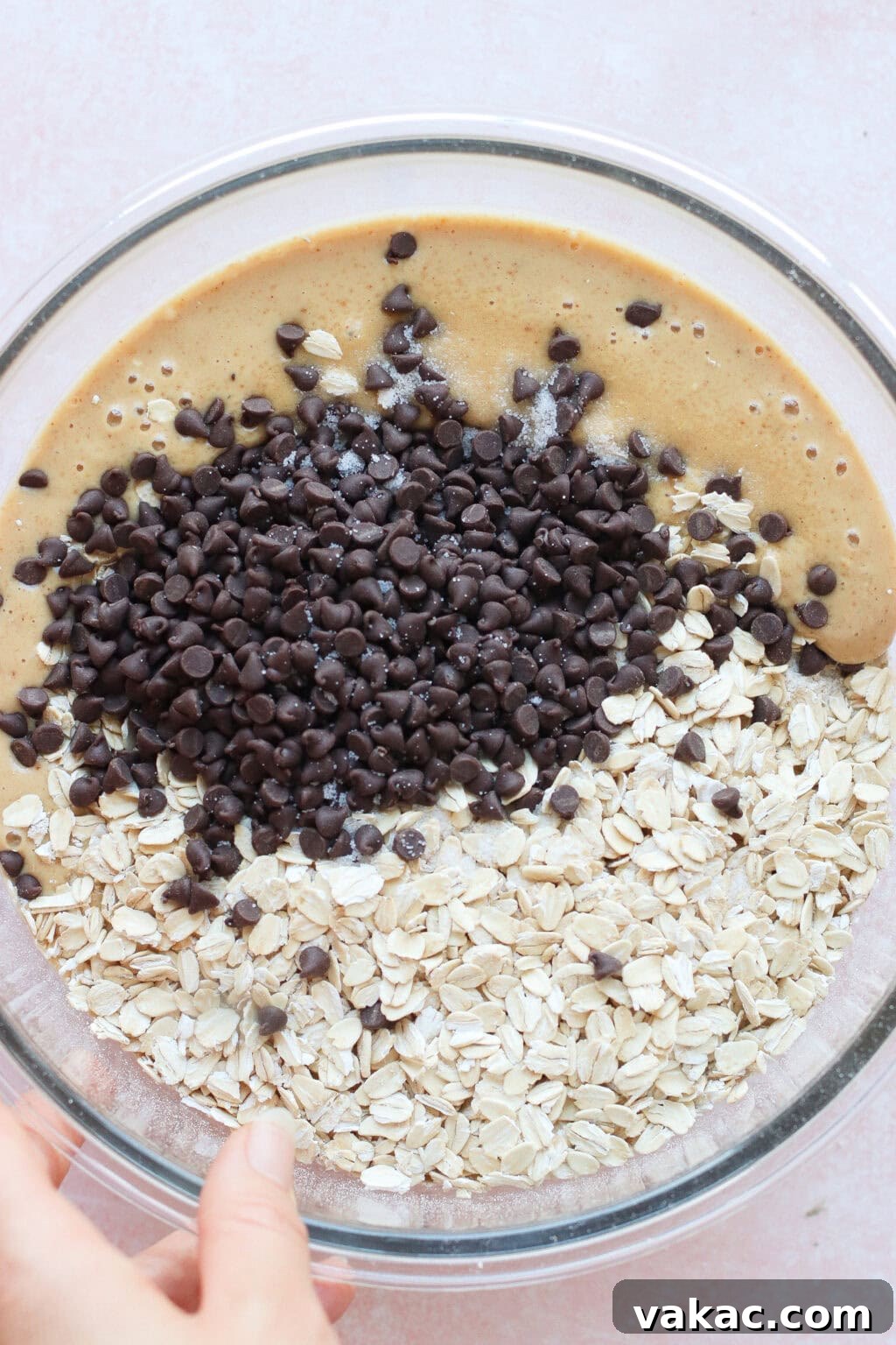 A glass bowl containing a mixture of oats, chocolate chips, and a light brown batter. A hand is holding the side of the bowl, preparing to mix the ingredients together.
