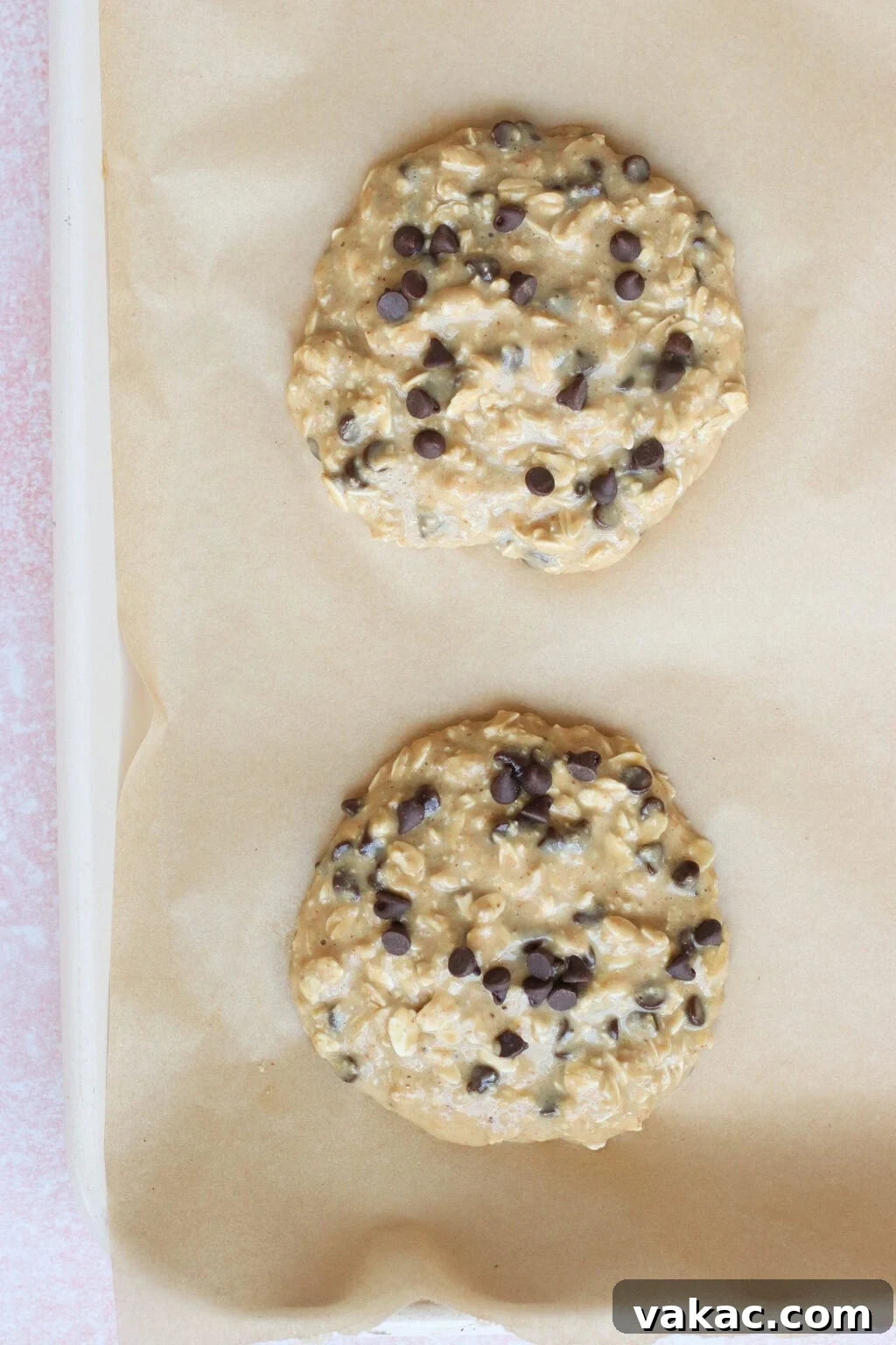 Two unbaked oatmeal chocolate chip cookies with visible chocolate chips sit on a parchment-lined baking sheet, ready to go into the oven.