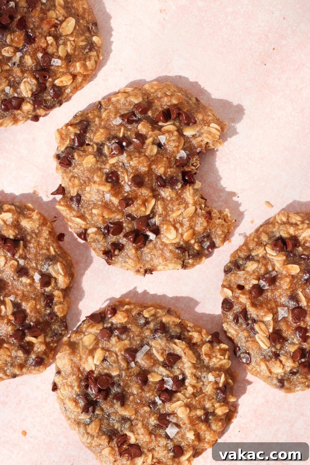 Five oatmeal chocolate chip cookies on a light pink surface, with one cookie missing a bite. The cookies are topped with chocolate chips and a sprinkle of flaky sea salt.