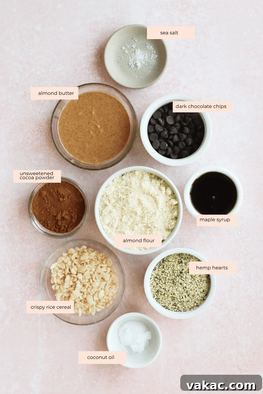 Chocolate Crunch Energy Balls Without Protein Powder 3 A flat lay of bowls containing sea salt, almond butter, dark chocolate chips, unsweetened cocoa powder, almond flour, maple syrup, crispy rice cereal, hemp hearts, and coconut oil on a light pink surface. Labels identify each ingredient.