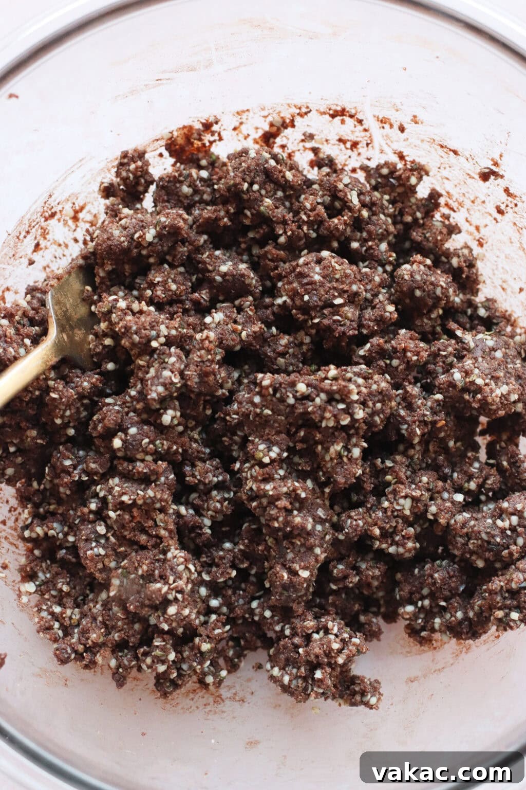 Chocolate Crunch Energy Balls Without Protein Powder 5 A close-up of a glass mixing bowl containing a thick, chunky mixture of chocolate and puffed grains, with a spoon partially visible on the left side.