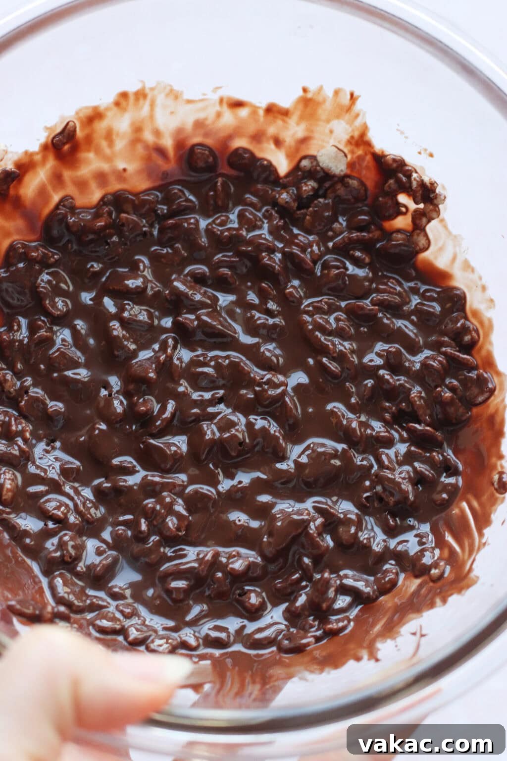Chocolate Crunch Energy Balls Without Protein Powder 7 A glass bowl filled with a mixture of melted chocolate and crispy rice cereal, partially stirred together, with some chocolate smeared along the sides of the bowl.