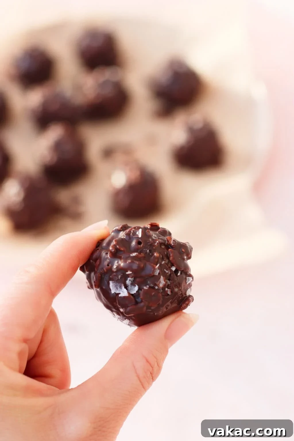 Chocolate Crunch Energy Balls Without Protein Powder 8 A hand holding a round, chocolate-covered treat with a textured surface; more similar treats are blurred in the background on parchment paper.