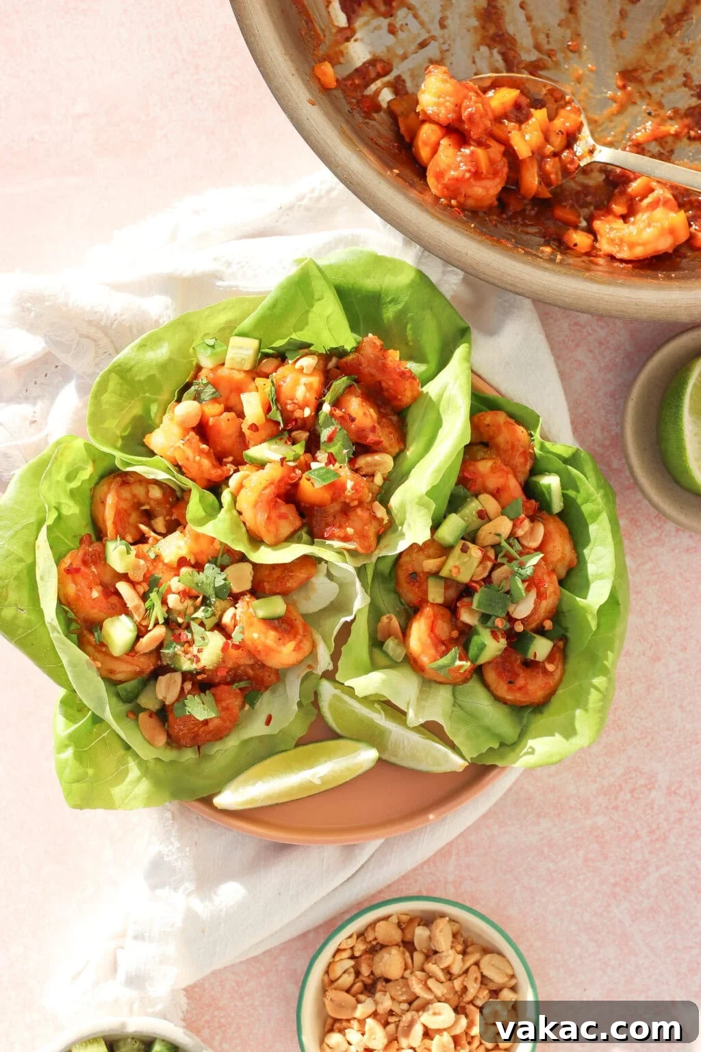 Honey-Soy Shrimp Lettuce Wraps with Crunchy Veggies 8 Three lettuce wraps filled with saucy shrimp, diced avocado, chopped peanuts, and herbs are arranged on a plate. A bowl of extra peanuts and a pan with remaining shrimp are nearby. Lime wedges are on the plate.