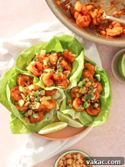 Three lettuce wraps filled with saucy shrimp, diced avocado, chopped peanuts, and herbs are arranged on a plate. A bowl of extra peanuts and a pan with remaining shrimp are nearby. Lime wedges are on the plate.
