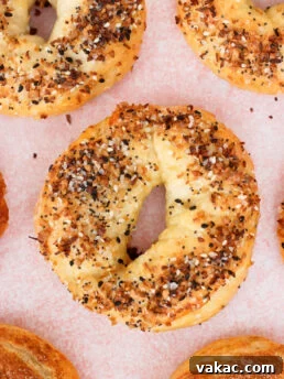A close-up of several everything bagels on a pink surface, topped with a mix of seeds and seasonings. The bagels are golden-brown and have a rough, homemade texture.