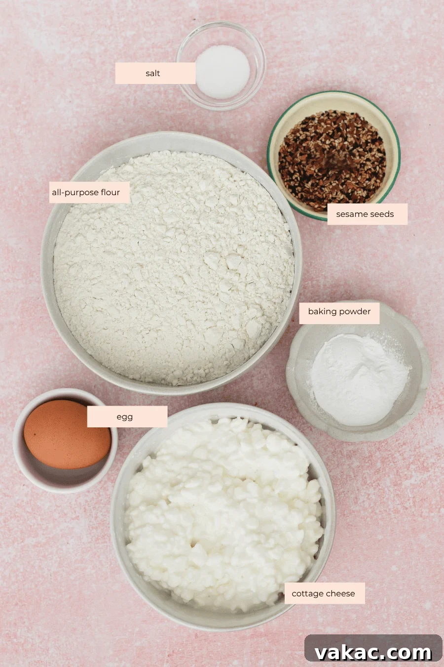 High-Protein Cottage Cheese Bagels Recipe for Homemade Bagels 3 Six labeled bowls on a pink surface containing salt, all-purpose flour, sesame seeds, baking powder, an egg, and cottage cheese, arranged neatly for a recipe preparation.